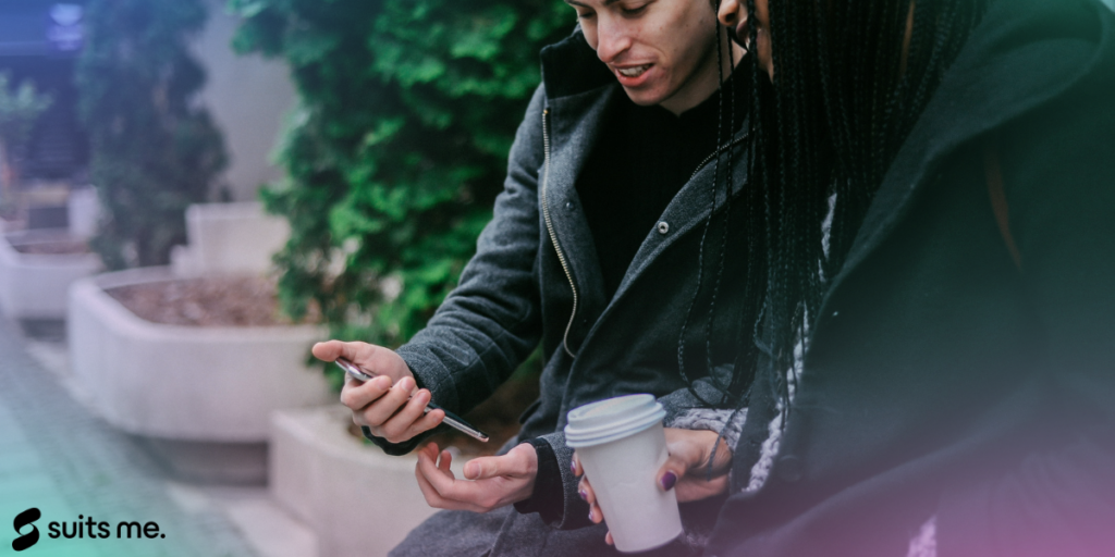 2 people sat together looking at a phone with a pink and green gradient and Suits Me logo in the bottom corner.