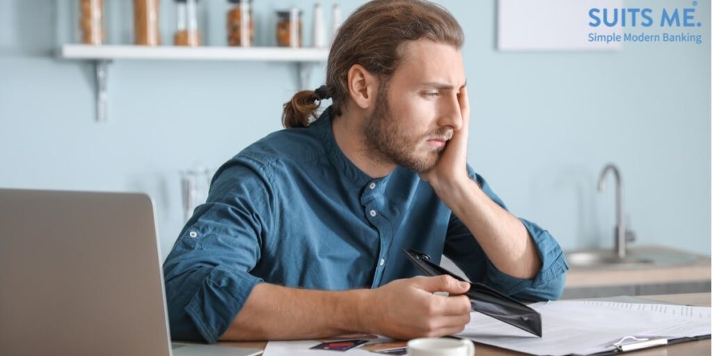 Young Man Sat in Kitchen Looks Worried, With No Money In His Wallet, Surrounded by debt letters, credit cards and laptop