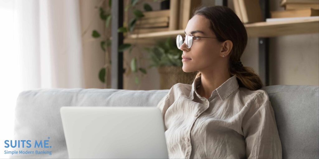 millennial woman sat on couch holding laptop look in distance thinking about money worries