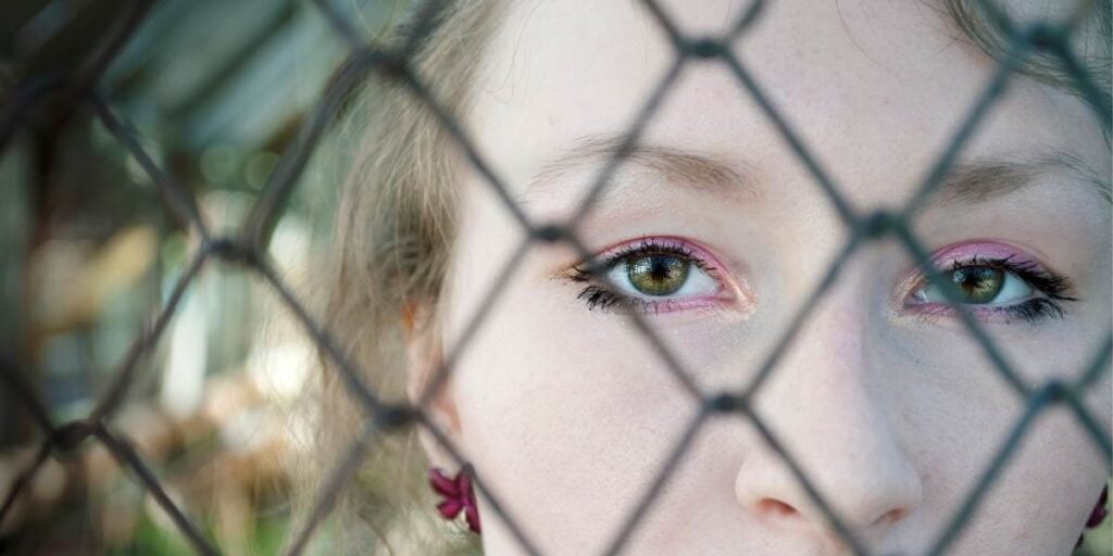 Image of a woman, with sadness in her eyes, behind a chain metal fence - representing being trapped within modern slavery and exploitation