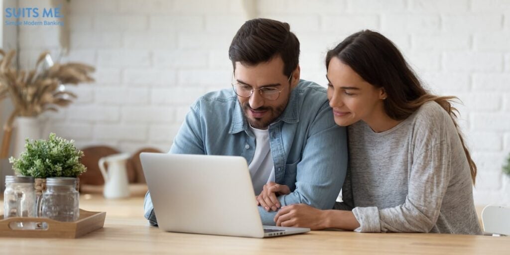 Couple sitting at big wooden table in modern kitchen, looking at laptop screen and managing their money though online banking