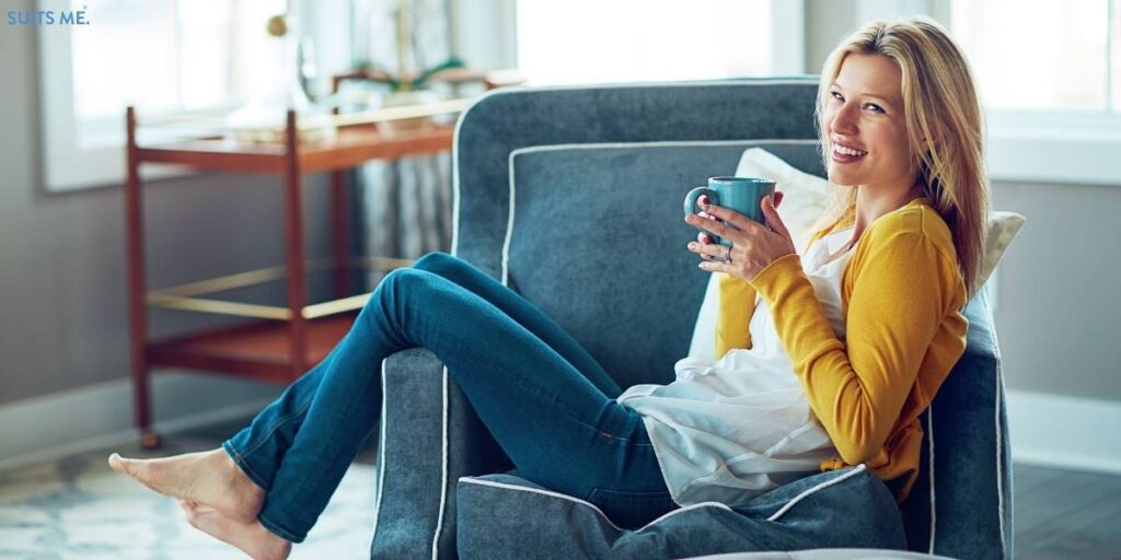 Happy woman sat over the arm of a blue chair, drinking a cup of tea and relaxing - representing unplugging from the world as a way to combat stress