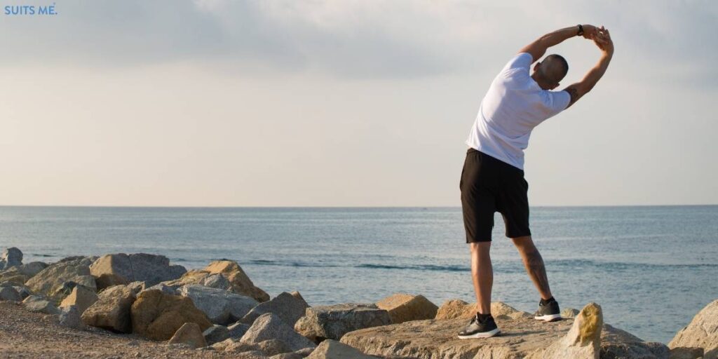 Image of man wearing activewear stretching and standing on a rock in front of the ocean - representing the importance exercising outdoors to alleviate stress.