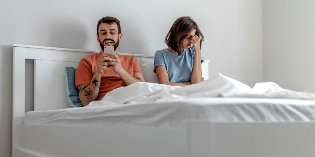 Couple in bed with the male laughing at his phone and the female looking upset signifying financial abuse