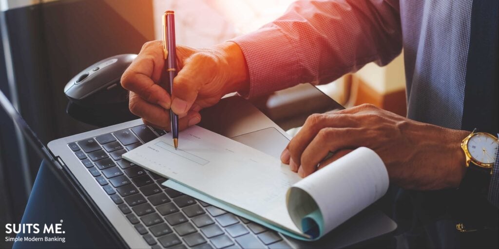 Businessman hand writing and signing white empty bank cheque to pay employees with modern computer notebook and wireless mouse on the desk at office. Payment by cheque, payroll concept